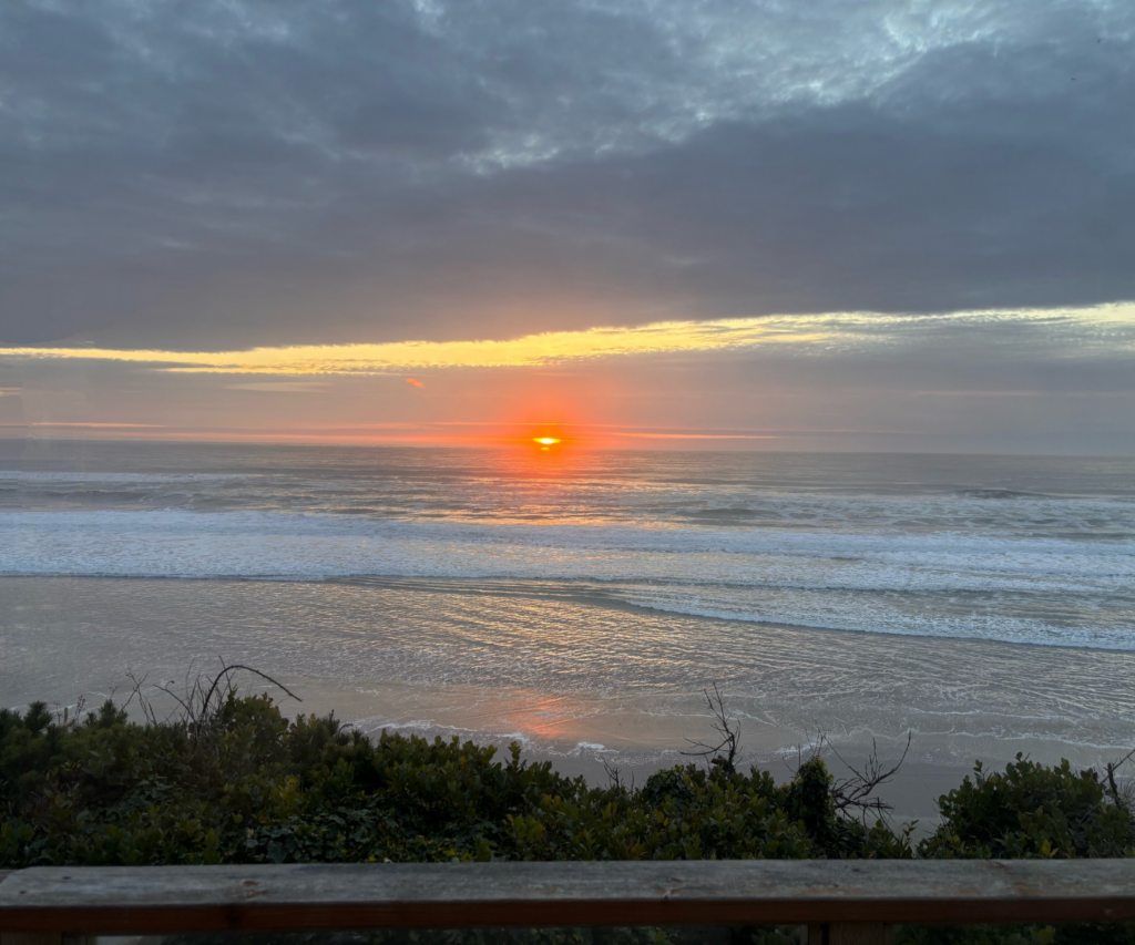 Bright orange sun setting over the pacific ocean with glowing clouds. With a deck and some bushes at the bottom.